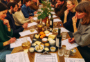 En livlig quizkveld med fokus på mat og drikke: folk rundt et bord med quizark, små smaksprøver, oste- og brødfat, vin- og kaffeprøver, varm stemning og små lapper med spørsmål